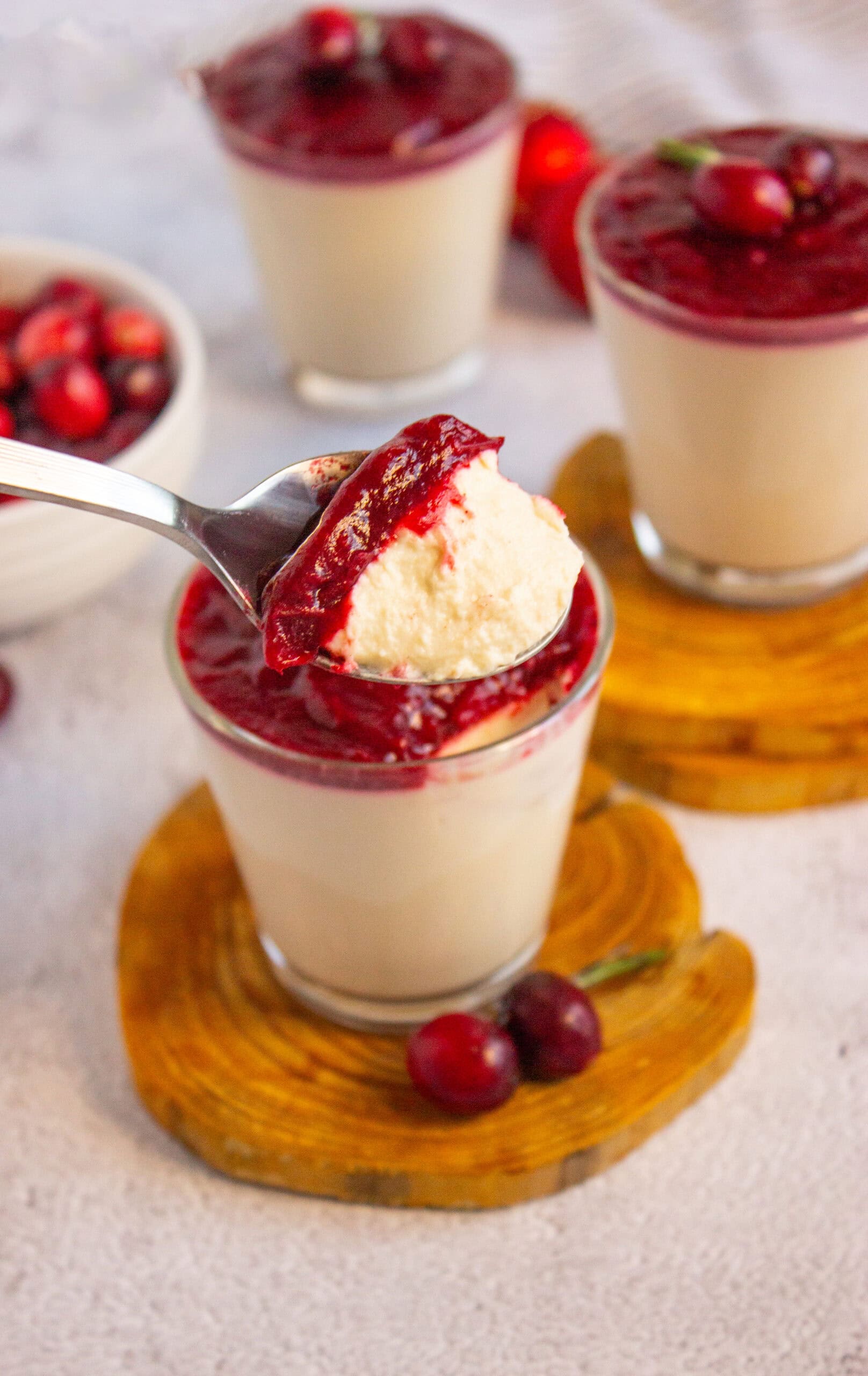 A spoonful of creamy Keto Cinnamon Panna Cotta with Cranberry Compote is held above a glass filled with the same dessert. Two more glasses and a bowl of cherries are in the background, all set on a light surface.