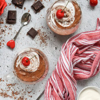 Two glass cups of chocolate mousse topped with whipped cream and cherries, surrounded by chocolate pieces, red roses, a red-and-white striped cloth, and a bowl of cream on a light textured surface.