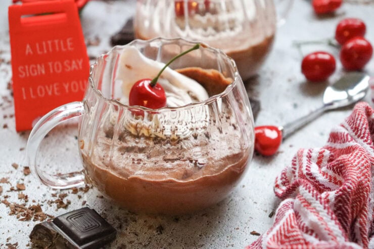 A glass cup of chocolate mousse topped with whipped cream and a cherry, next to a red cloth, chocolate pieces, fresh cherries, and a red sign that reads A little sign to say I love you.
