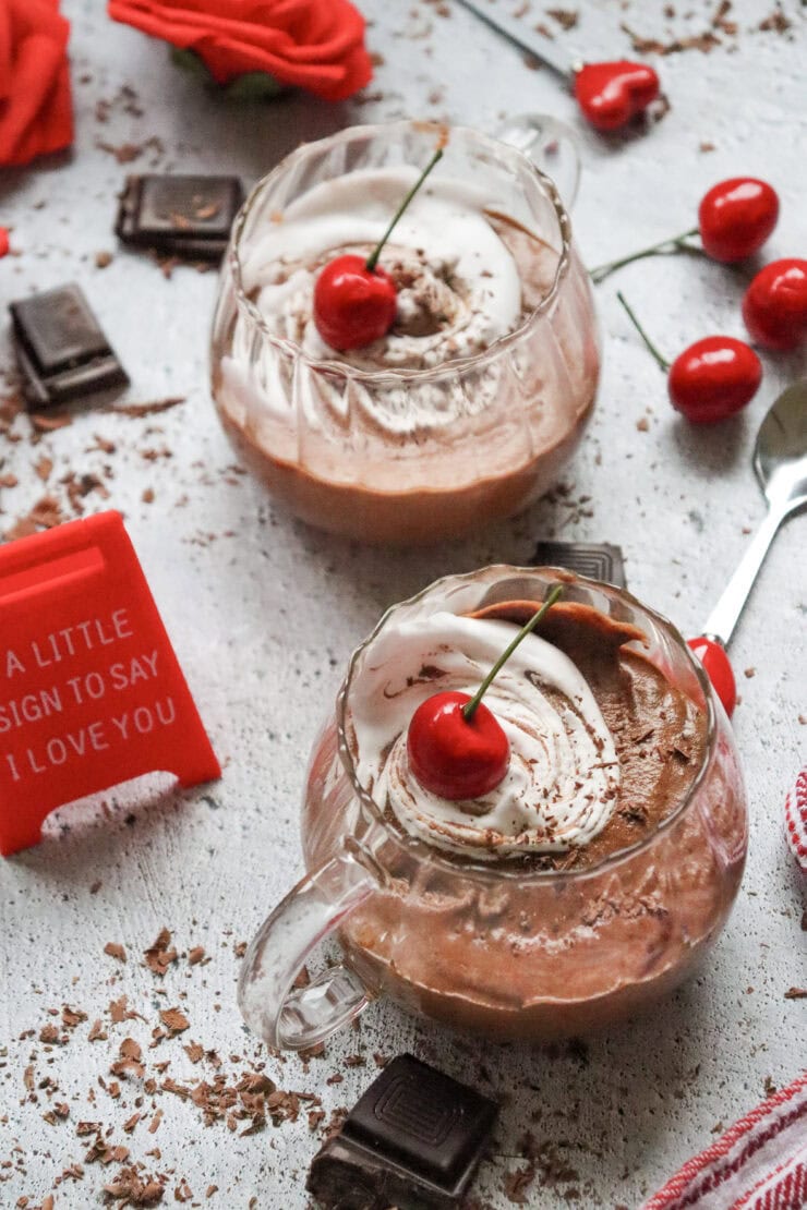 Two glass cups of creamy hot chocolate topped with whipped cream and cherries, surrounded by chocolate pieces, fresh cherries, a spoon, and a red sign that says, A little sign to say I love you.