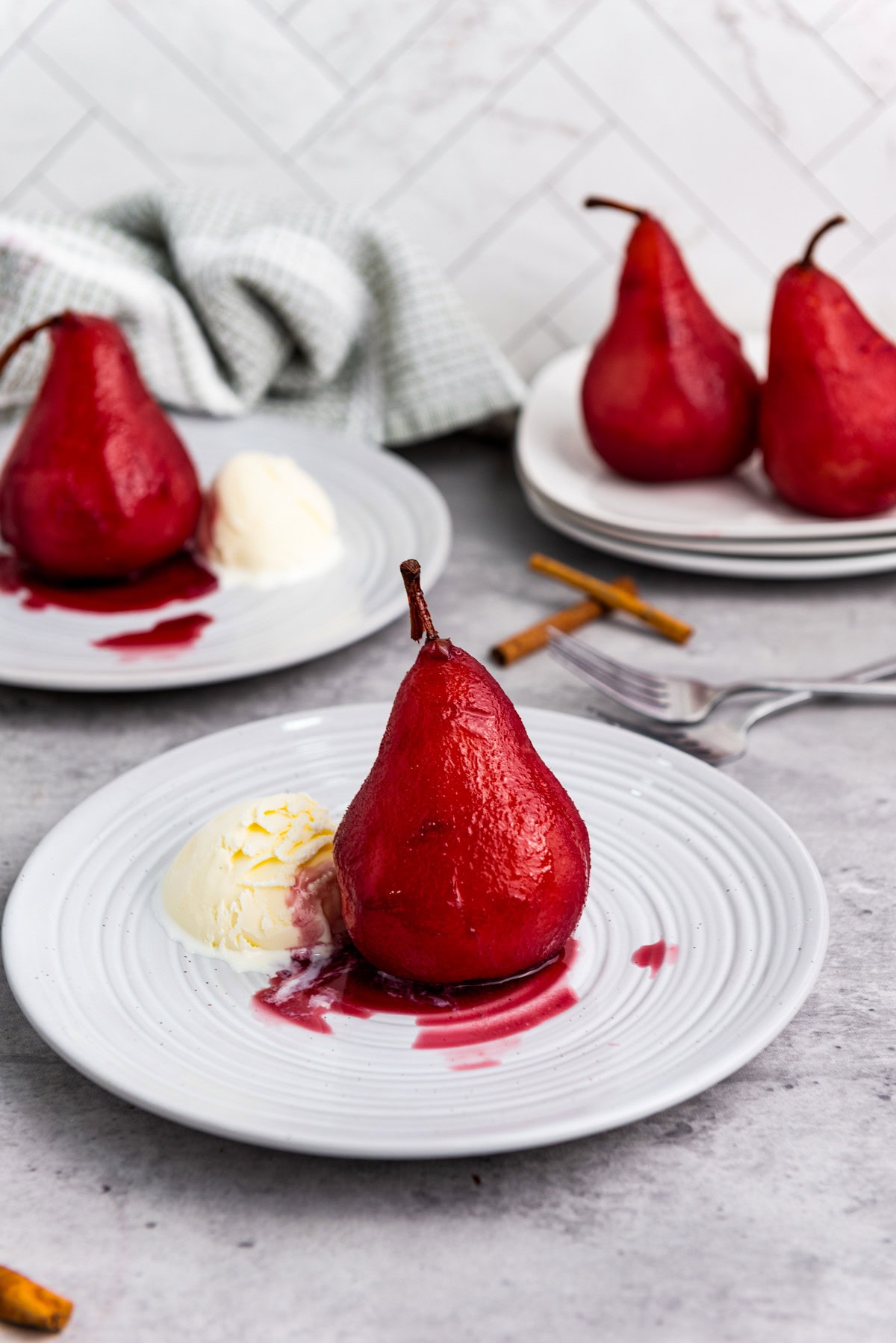 A poached pear in red wine sauce sits on a white plate with a scoop of vanilla ice cream. In the background, more poached pears are on another plate, with a fork and a towel nearby.