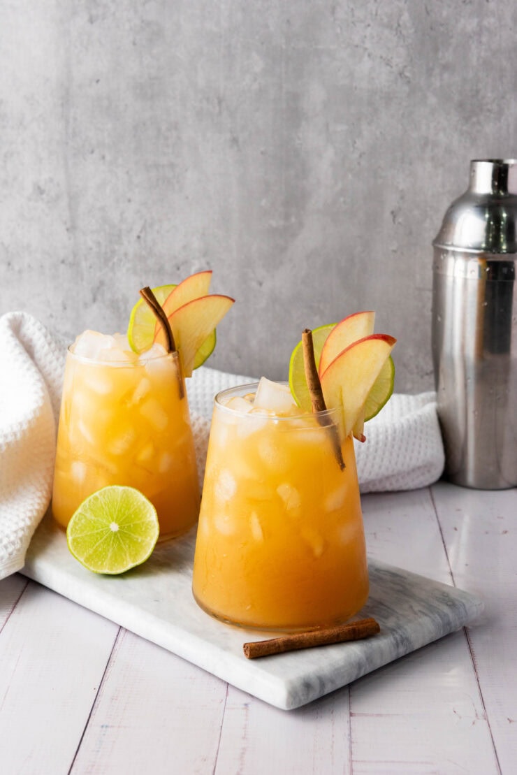Two iced cocktails garnished with apple slices, cinnamon sticks, and lime sit on a marble tray. A lime half and a cocktail shaker are nearby, with a white towel in the background.
