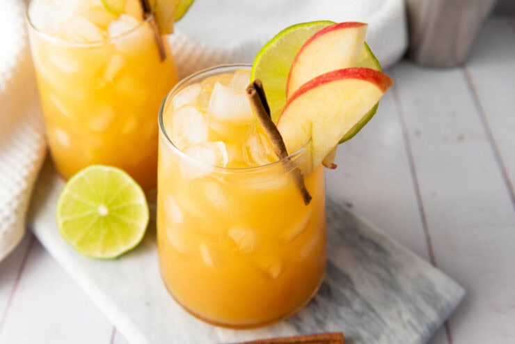 A glass of iced orange drink garnished with apple slices, a lime wheel, and a cinnamon stick, sits on a marble board, with a lime half and another drink in the background.