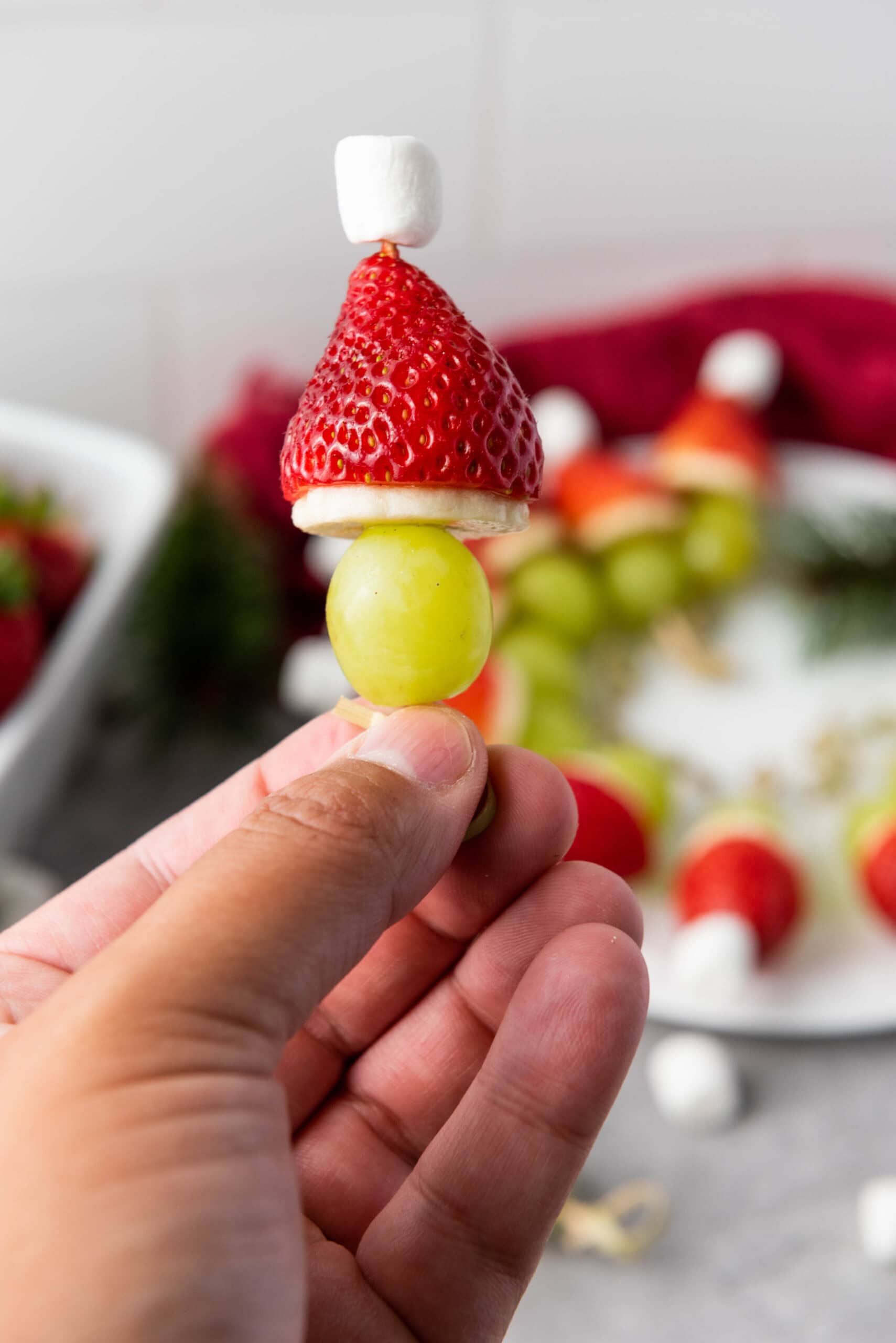 A hand holds a festive snack made of a green grape, a slice of banana, a strawberry, and a mini marshmallow stacked on a toothpick, resembling a holiday gnome or Santa. A plate with similar snacks is in the background.