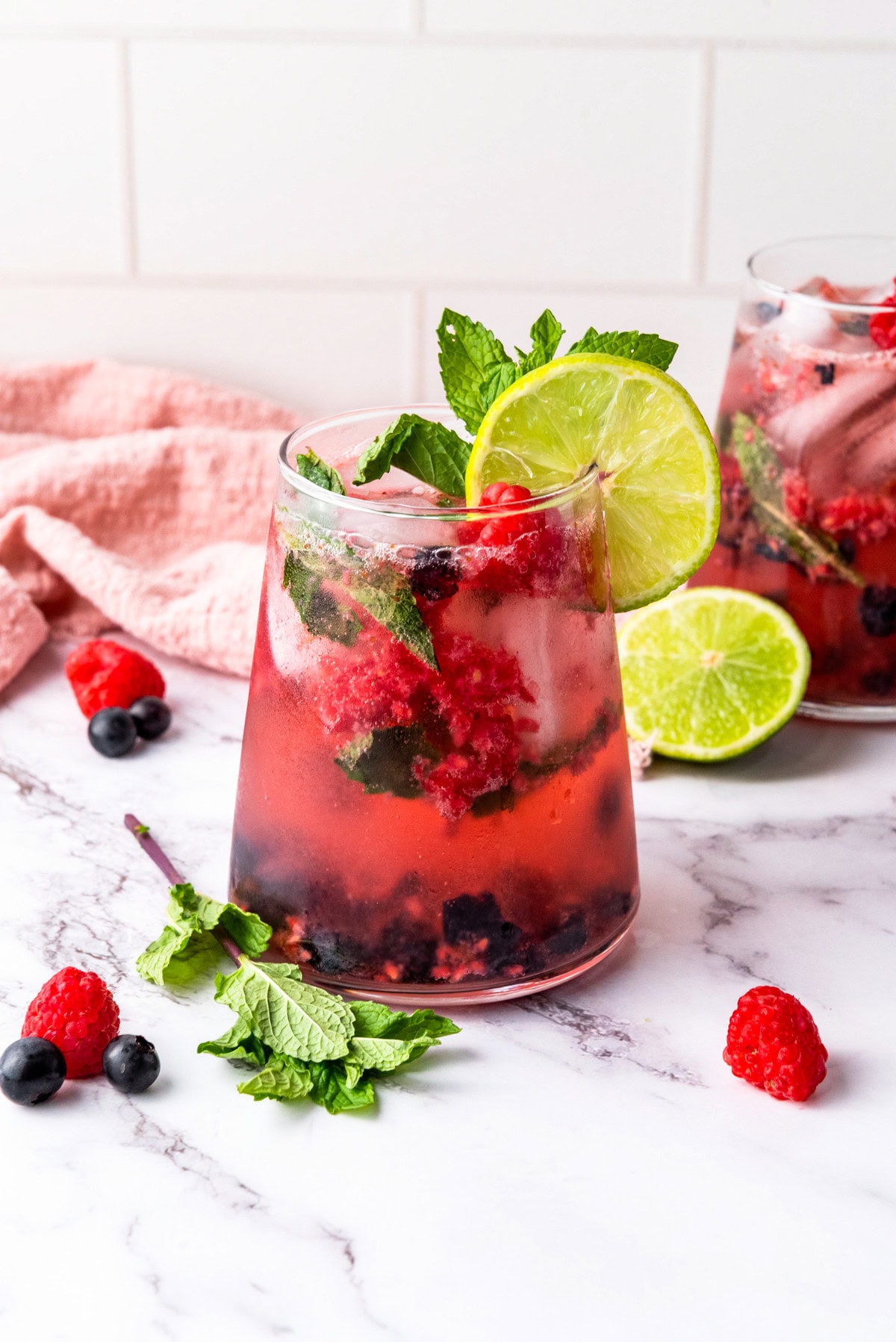 A glass of berry mojito garnished with lime, mint, raspberries, and blueberries on a marble surface, with a second glass, lime slices, and a pink towel in the background.