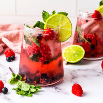 Two glasses of Summer Berry Cooler with ice, mint leaves, lime slices, raspberries, and blueberries on a marble surface. A pink cloth and loose berries decorate the background.