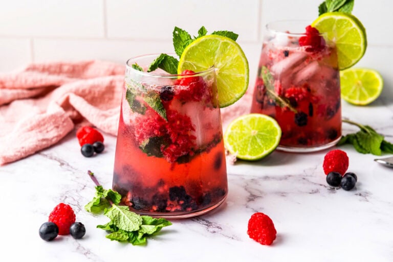 Two glasses of Summer Berry Cooler with ice, mint leaves, lime slices, raspberries, and blueberries on a marble surface. A pink cloth and loose berries decorate the background.