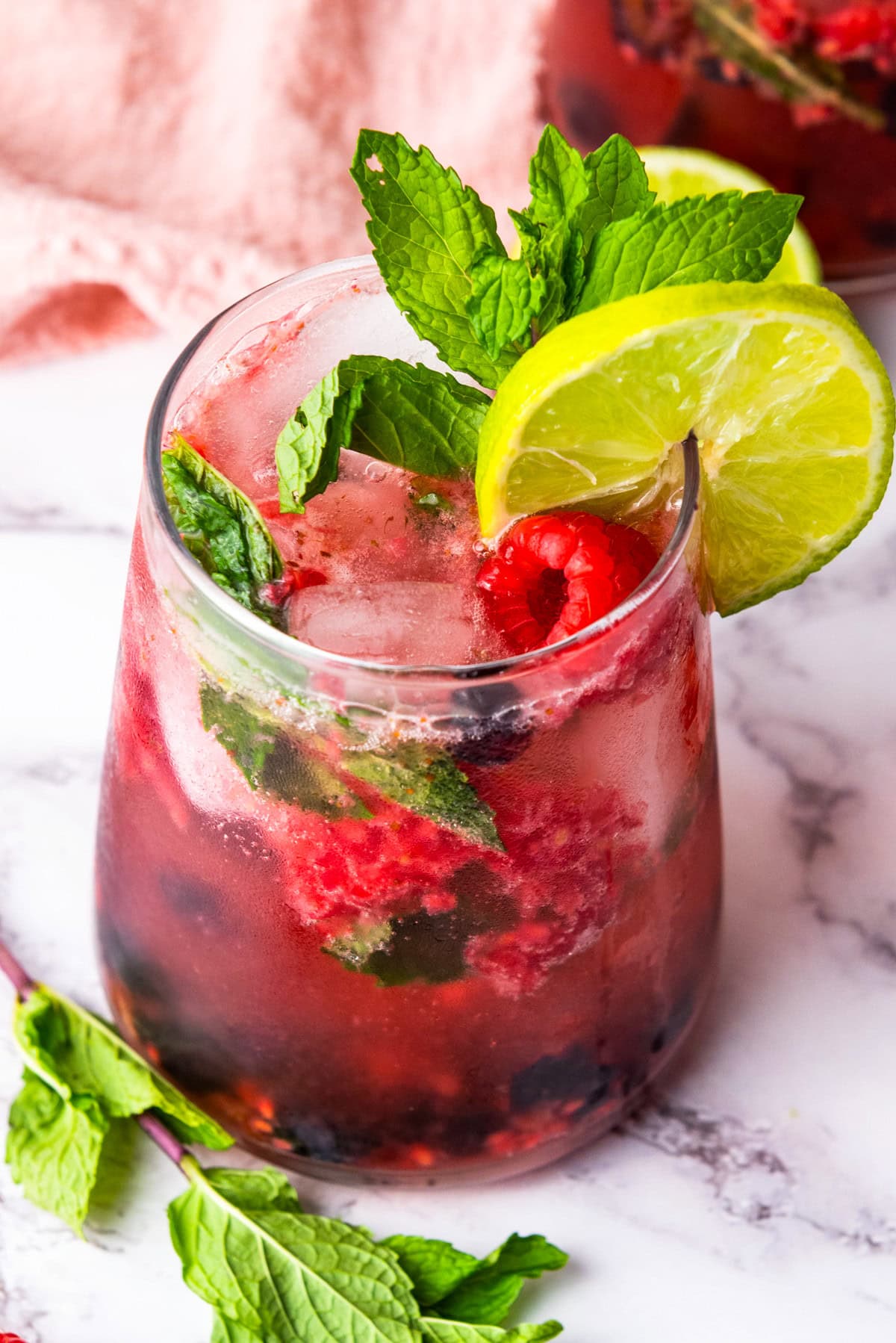 A glass of berry mojito with ice, fresh mint, raspberries, and a lime slice garnish, placed on a white marble surface with mint leaves nearby.