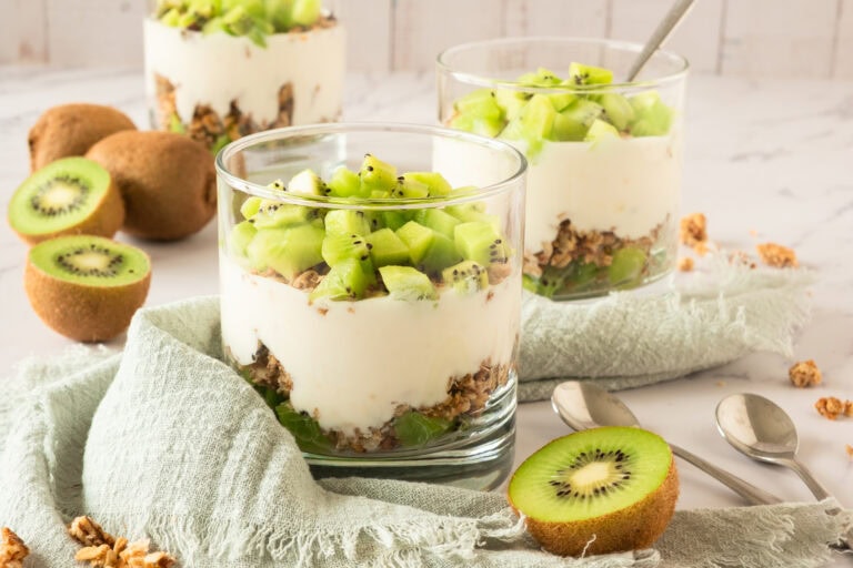 Three glass cups filled with layers of granola, yogurt, and chopped kiwi sit on a light green cloth. Sliced and whole kiwis, spoons, and extra granola are arranged around the cups on a white surface.