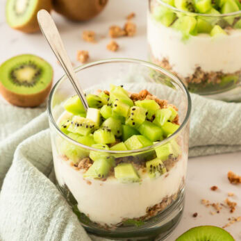 A glass filled with layers of yogurt, granola, and diced kiwi, topped with a spoon. Whole and sliced kiwis are placed nearby on a light green cloth, with a second parfait glass in the background.
