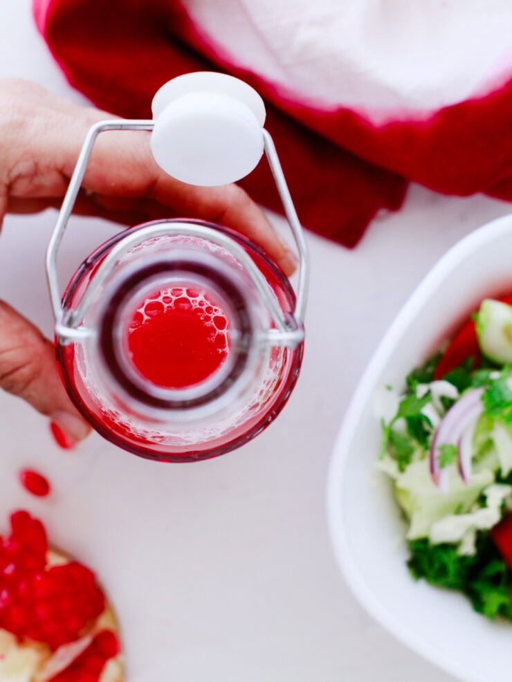 A hand holds a glass bottle of red liquid with a white flip-top cap, seen from above. Next to it are a bowl of fresh salad and a red cloth on a white surface.