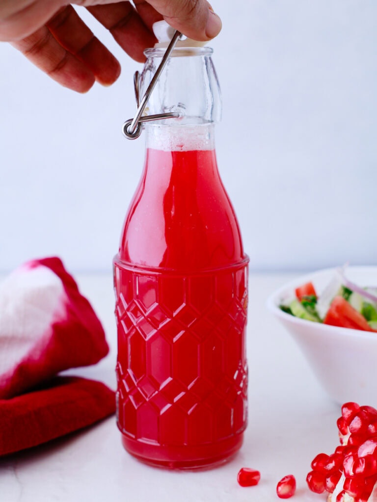 A hand holds a glass bottle filled with bright red juice, featuring a metal clasp lid. Nearby are a red cloth, some pomegranate seeds, and a salad bowl on a white surface.