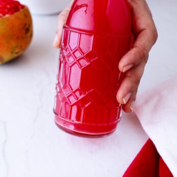 A hand holding a patterned glass bottle filled with bright red juice. Pomegranate seeds and a pomegranate are on the white surface, with a red cloth and a white bowl in the background.