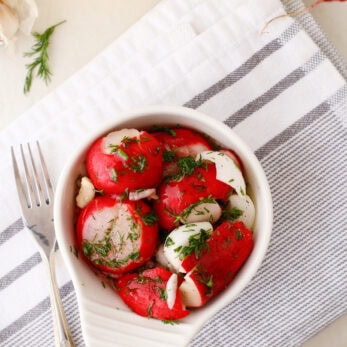 A white bowl of sliced radishes fresh dill sits on a striped kitchen towel, next to a fork, garlic, herbs, and a bottle of olive oil.