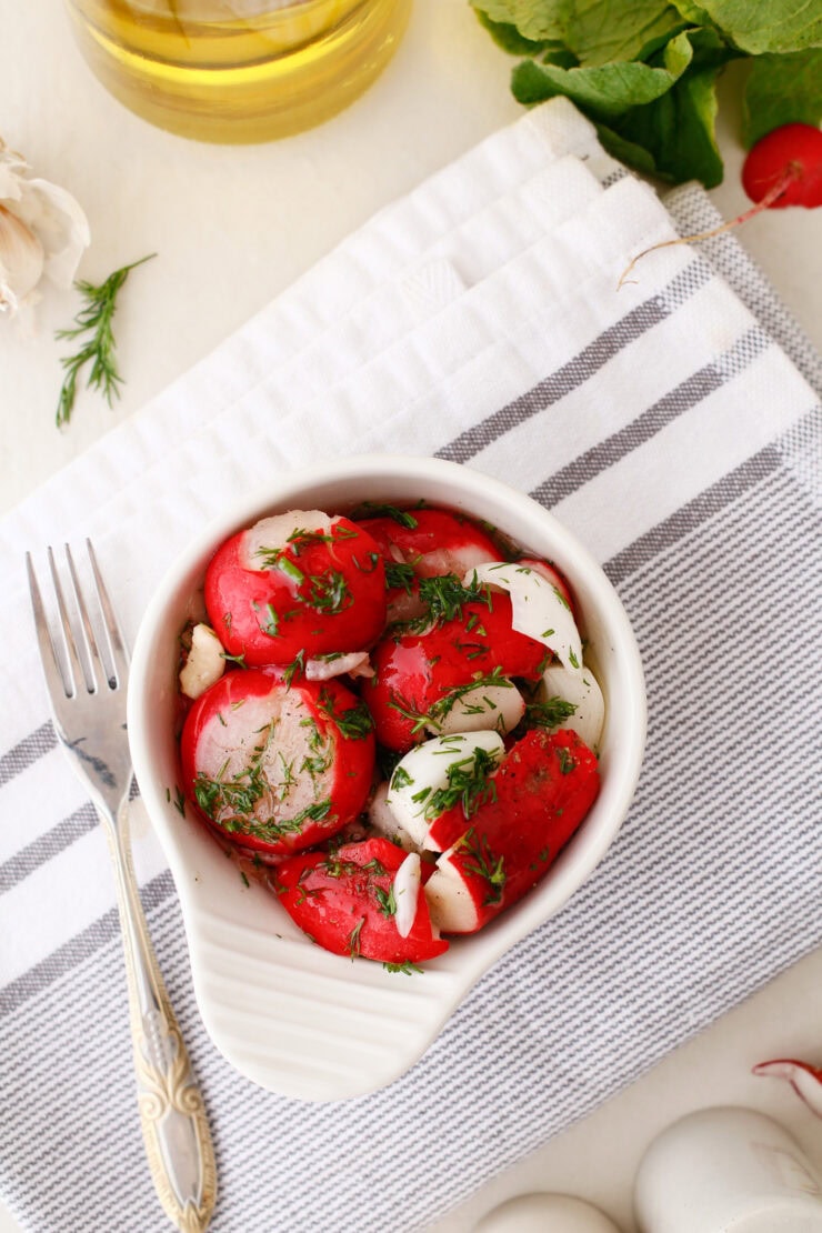 A white bowl of sliced radishes and mozzarella balls garnished with fresh dill sits on a striped kitchen towel, next to a fork, garlic, herbs, and a bottle of olive oil.