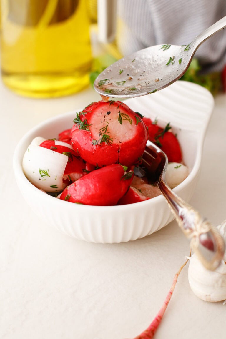 A white bowl filled with sliced radishes and chopped herbs. A spoon above the bowl is being used to drizzle dressing or sauce over the radishes. A fork rests in the bowl, and an olive oil bottle is in the background.