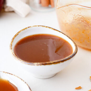 A small, white, rustic bowl filled with smooth apple molasses l sauce sits on a white surface, with a plate holding a spoonful of sauce and a glass bowl with remnants of sauce nearby.
