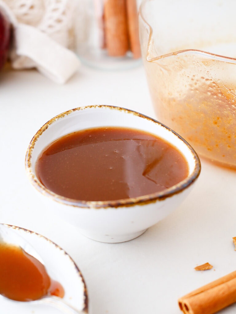 A small, white, rustic bowl filled with smooth apple molasses l sauce sits on a white surface, with a plate holding a spoonful of sauce and a glass bowl with remnants of sauce nearby.