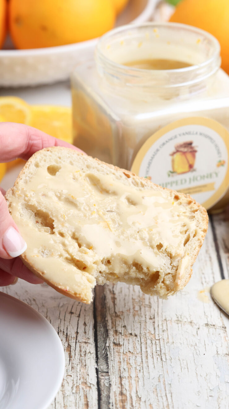 A hand holding a slice of bread spread with whipped honey, with a jar of the same honey in the background.