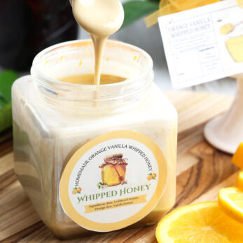 A spoonful of cream-colored whipped honey being drizzled into a square glass jar labeled Homemade Orange Vanilla Whipped Honey, set on a wooden board next to orange slices.