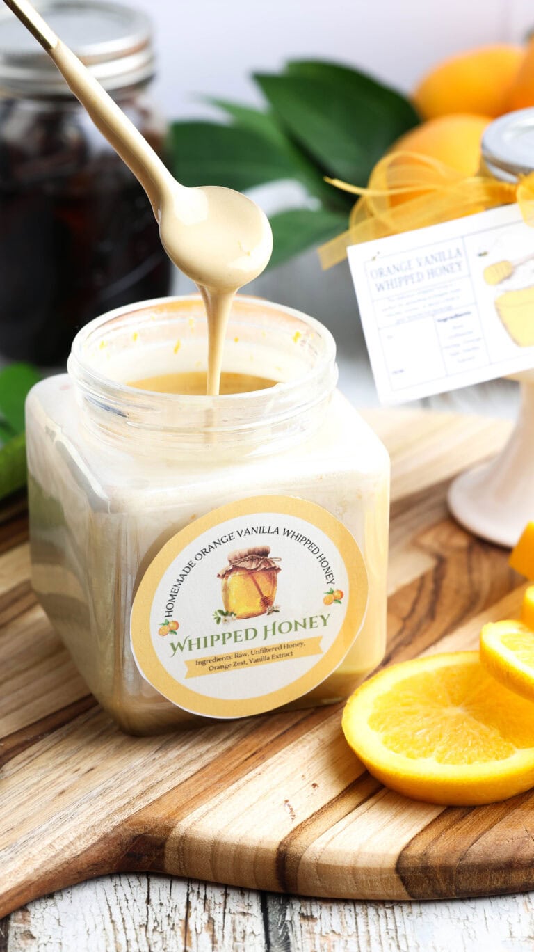 A spoonful of cream-colored whipped honey being drizzled into a square glass jar labeled Homemade Orange Vanilla Whipped Honey, set on a wooden board next to orange slices.