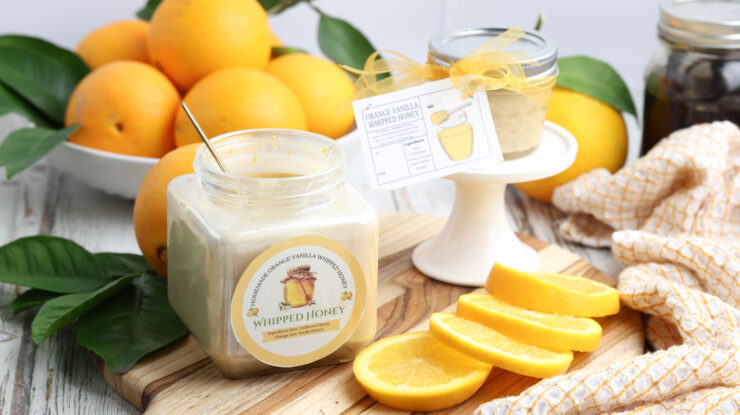 Jars of orange vanilla whipped honey with labels on a wooden board, surrounded by fresh oranges and slices.