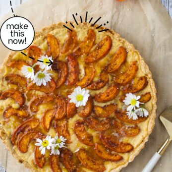 Top view of a golden apricot tart with custard, decorated with white flowers and placed on parchment paper beside a cake server. Text overlay reads “Apricot Tart with Custard (Gluten-Free).”.
