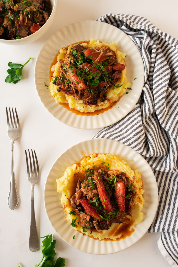 Two plates of mashed potatoes topped with shredded beef, carrots, and herbs, with forks and a striped napkin on the side.