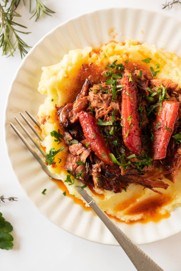 Braised beef and carrots in gravy served over a mound of mashed potatoes on a white plate with a fork.