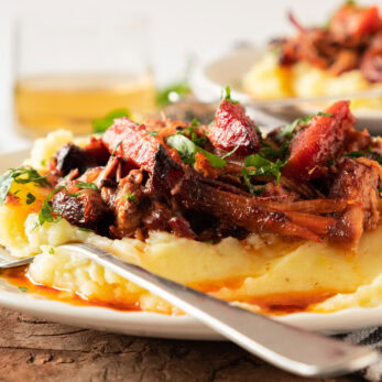 Slow-Cooked Pork Pot Roast topped with fresh herbs served over a bed of mashed potatoes on a white plate with a fork.