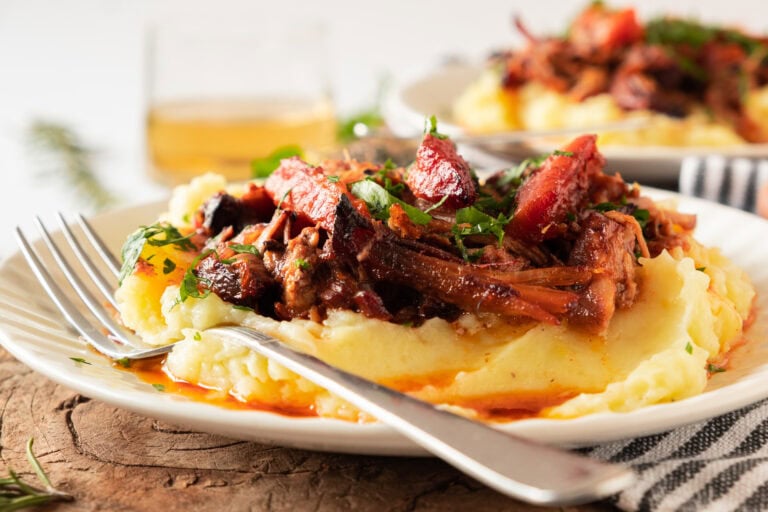 Slow-Cooked Pork Pot Roast topped with fresh herbs served over a bed of mashed potatoes on a white plate with a fork.