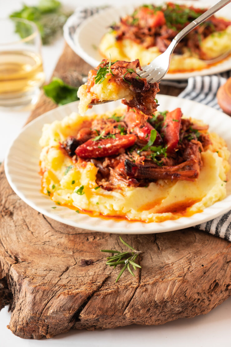 A fork lifts a bite of shredded meat and vegetables over a plate of mashed potatoes.
