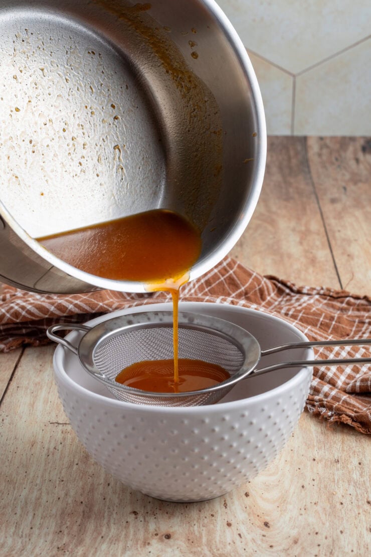 A pot of orange-brown liquid is being poured through a metal strainer into a white textured bowl on a wooden surface, with a brown checkered cloth in the background.