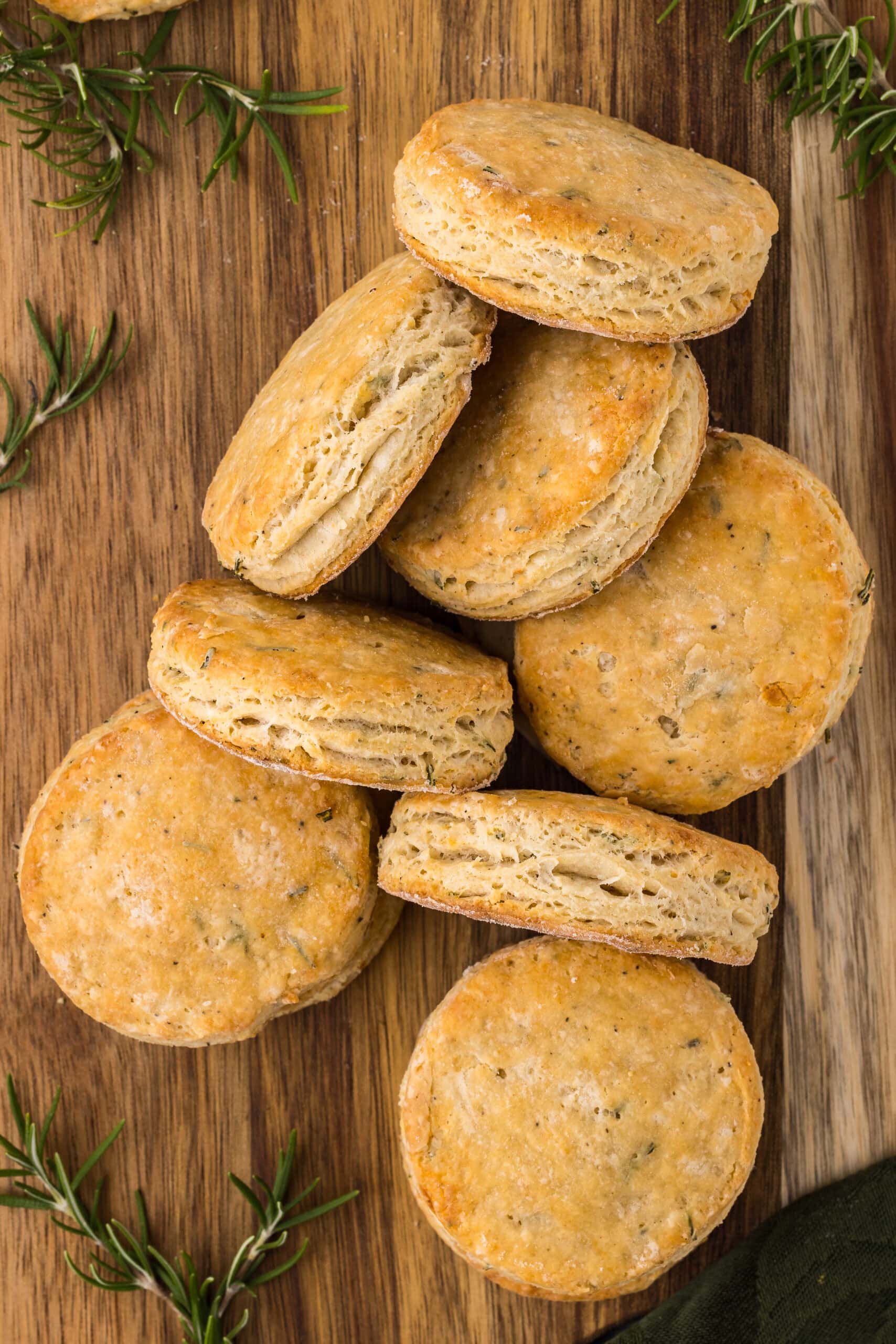 Round, golden-brown gluten-free biscuits are stacked and scattered on a wooden surface, surrounded by sprigs of fresh rosemary. The biscuits appear flaky and freshly baked.