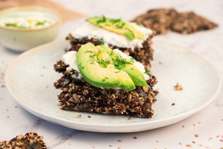 Two slices of seed crackers topped with cream cheese, avocado slices, and fresh herbs are on a white plate. There is a small dish with dip in the background on a marble surface.