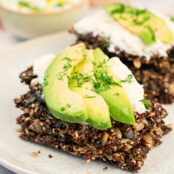 Seed crackers topped with sliced avocado, a dollop of cream, and fresh herbs are stacked on a plate. A bowl of dip is blurred in the background. The dish appears healthy and appetizing.