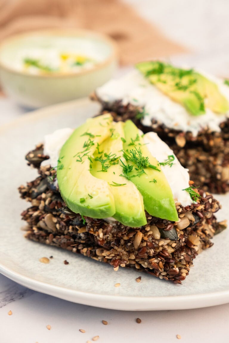 Seed crackers topped with sliced avocado, a dollop of cream, and fresh herbs are stacked on a plate. A bowl of dip is blurred in the background. The dish appears healthy and appetizing.