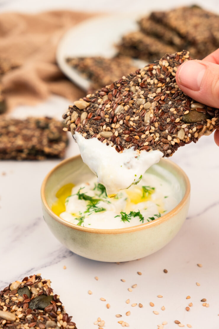 A hand dips a seeded cracker into a small bowl of creamy white dip garnished with olive oil and fresh herbs. More crackers and a plate are visible in the background on a light surface.