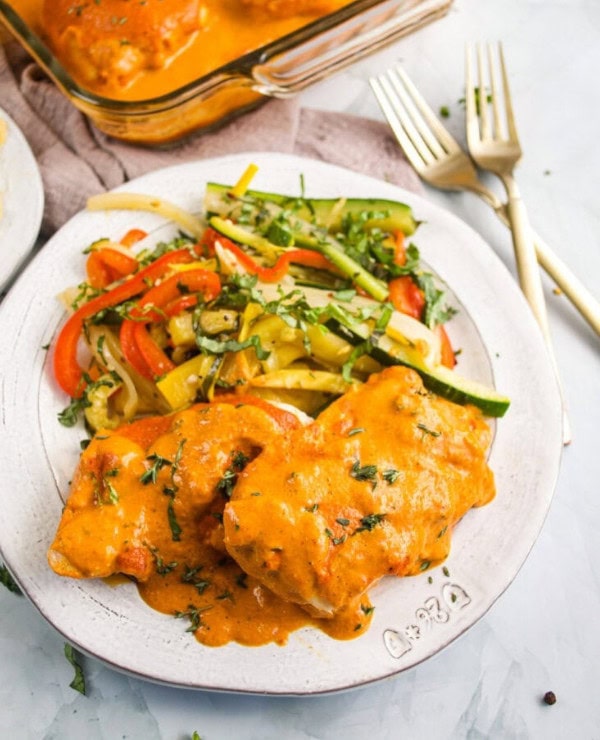 A white plate with two pieces of chicken covered in creamy orange sauce, served alongside sautéed vegetables including zucchini, bell peppers, and yellow squash. A glass baking dish and two forks are in the background.