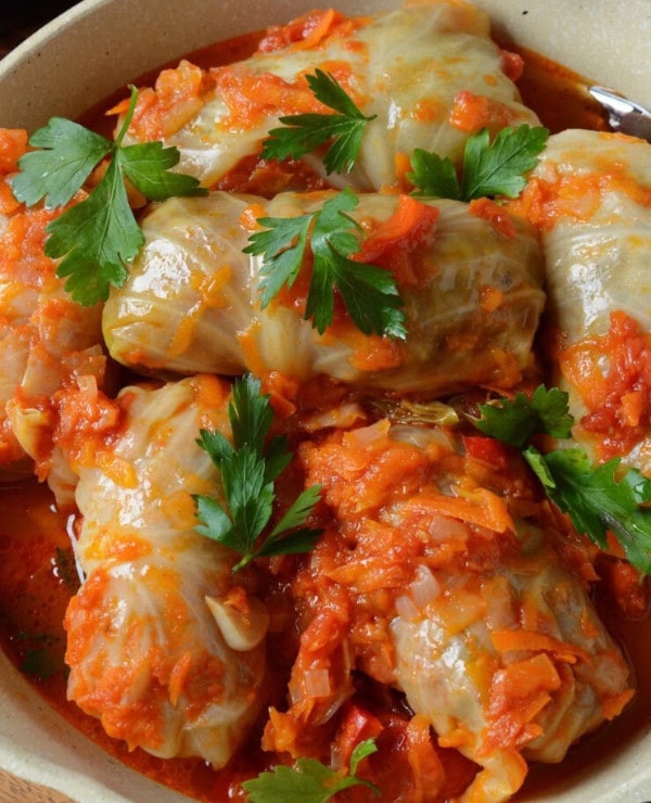 A bowl of cabbage rolls stuffed with filling, topped with a chunky tomato and carrot sauce, and garnished with fresh parsley leaves.