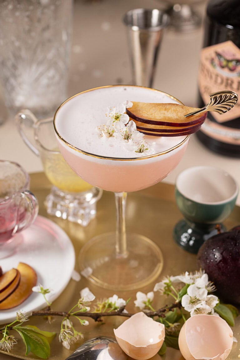 Elegant pink gin cocktail in a vintage coupe glass, topped with a frothy layer of foam, delicate white blossoms, and thin slices of plum on a decorative pick. Surrounding the drink are ingredients and props including halved plums, an eggshell, floral branch, jiggers, and a dark liquor bottle, creating a stylish, spring-inspired mixology scene.