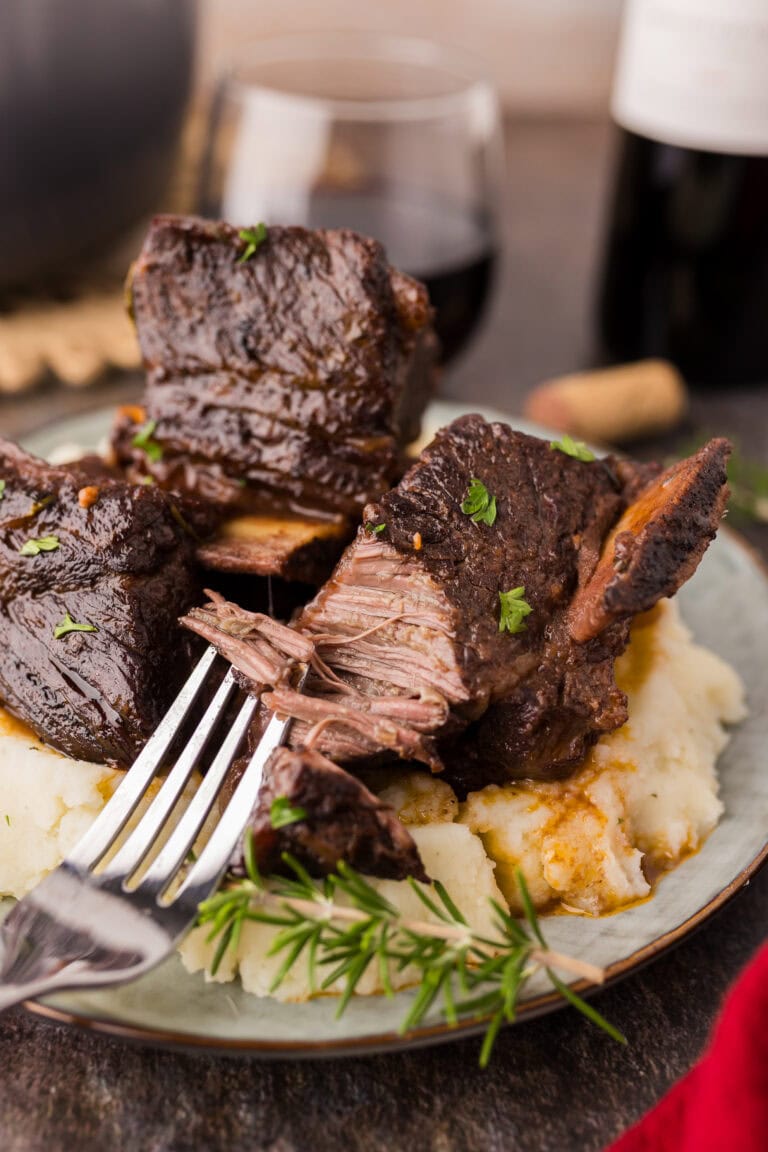 Close-up of tender red wine braised short ribs served in a rich, glossy sauce with carrots and fresh herbs, plated in a rustic dish over a light stone background.